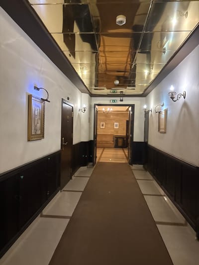 This is an eye-level, slightly low-angle photograph capturing a long, ornate interior hallway located in Kyustendil, Bulgaria. The perspective is directed down the length of the hallway towards an open doorway at the far end.
The ceiling is covered with multiple rectangular, gold-toned, reflective panels, creating a mirrored effect that reflects the floor, walls, and lighting fixtures. A circular white smoke detector is visible mounted on the ceiling near the center.
The walls feature a two-tone design: the upper section is painted white, while the lower section consists of dark brown wooden wainscoting or paneling. Several decorative elements are affixed to the walls. On both sides, antique-style sconce lights with exposed flame-shaped bulbs cast a warm glow. Above framed artworks, modern picture lights emit a distinct blue-white illumination, highlighting the artwork below. A rectangular framed picture with a gold-colored border is prominently displayed on the left wall, illuminated by a blue-tinted picture light. A similar but smaller framed picture is visible on the right wall.
Multiple dark brown wooden doors, possibly leading to rooms or other areas, are spaced along both sides of the hallway. Each door features a circular gold-colored escutcheon and a matching handle. One door on the left is fully closed, while the doorway at the far end of the hallway is wide open, revealing an interior room with a sofa, a table, and additional framed pictures on its walls. Through this open doorway, two green and white emergency exit signs are visible, one pointing left and one indicating straight ahead. The room beyond appears to have a different flooring, possibly wooden planks, and features a chandelier in the distance.
The floor of the hallway consists of a long, dark brown carpet runner extending down the center. This carpet is flanked on both sides by lighter-colored, rectangular floor tiles or marble strips. No individuals or active interactions are present in the scene.