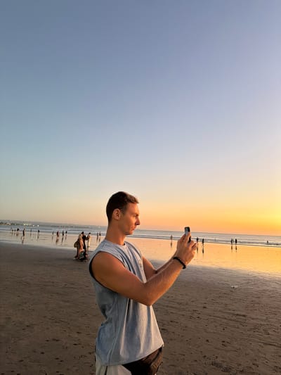 A man stands on the wet sand of Kuta Beach, Indonesia, holding a smartphone horizontally to take a photograph. He is wearing a light blue sleeveless t-shirt and dark shorts. His left arm is extended, holding the phone, and his right hand is on the side of the phone, also holding it. He is looking towards the phone, his profile visible. In the background, the ocean stretches to the horizon, with a warm sunset casting an orange glow across the sky and reflecting on the wet sand and water. Several other people are scattered across the beach, some walking, others standing, their figures silhouetted against the vibrant sunset. The sky transitions from a pale blue at the top to a rich orange near the horizon.
