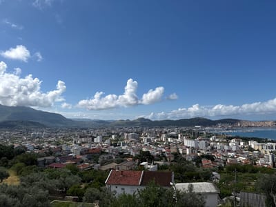 This panoramic photograph captures an expansive daytime view of the coastal town of Šušanj, Montenegro, under a bright blue sky dotted with scattered white cumulus clouds. The perspective is from an elevated position, looking down over the urban landscape towards the sea and distant mountains.

In the immediate foreground, dense green foliage, including what appear to be olive groves, frames the bottom of the image. A prominent white house with a reddish-brown tiled roof stands out among the trees.

The midground is dominated by the sprawling town. Numerous buildings, a mix of residential houses with terracotta roofs and taller, more modern apartment blocks, extend across the scene. The architecture suggests a blend of traditional and contemporary styles. Green spaces and trees are interspersed throughout the urban fabric.

Further into the background, a large body of water, likely a bay or the Adriatic Sea, is visible on the right, featuring a marina with several boats. The coastline is rugged, with some exposed rock faces. Majestic green mountains and hills rise behind the town, extending across the horizon. On some distant ridges, subtle indications of wind turbines can be seen.

The scene depicts a sunny day with pleasant weather conditions, likely morning or early afternoon given the bright light. No specific human activity or visible text is discernible. The overall impression is one of a vibrant coastal settlement nestled between mountains and the sea.