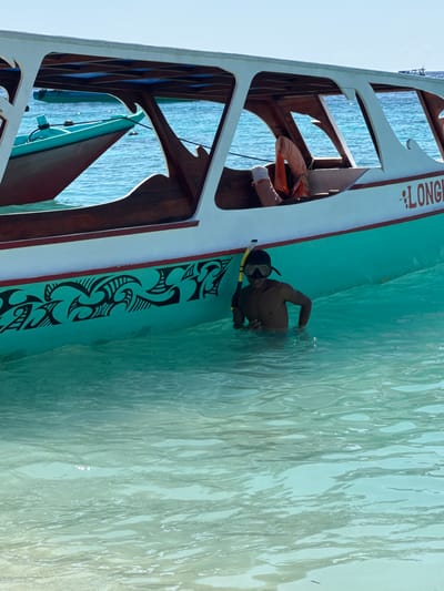 An unidentified individual, wearing a snorkel mask and holding a snorkel, stands in shallow, clear turquoise water near a boat. The boat is partially submerged in the water and has a white exterior with teal accents and a wooden interior. A decorative tribal pattern in black is visible on the teal portion of the boat's hull. The boat has an orange life vest visible inside. Another boat, teal and maroon, is tied to the side of the main boat. The background shows a bright blue sky and the ocean. The location is stated as Unknown, Indonesia.