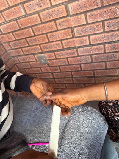 The image shows a close-up of two people's hands during a nail filing session. The background features a textured brick wall in shades of red and brown, with a silver electrical outlet plate visible. One person's hand, adorned with a silver bracelet, is positioned on the left side of the frame, with their nails appearing to be in the process of being filed. The other person's hands are on the right, holding a white nail file and working on the fingernails. This individual is wearing a grey towel or robe. The scene appears to be indoors, possibly in a salon or home setting in Ajara, Nigeria. The lighting suggests daytime. No visible text is present in the image.