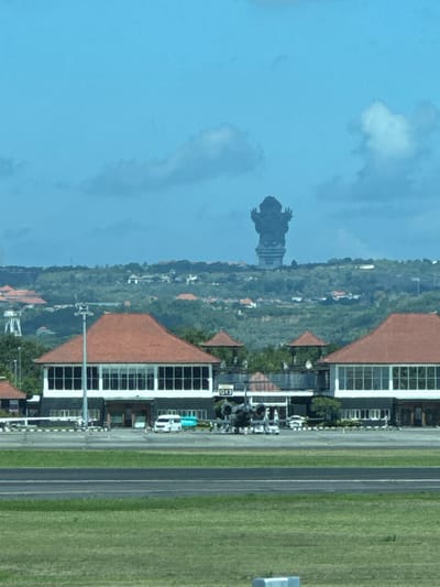 The image captures a wide-angle view of an airport tarmac with a large, dark statue visible on a distant hill. In the foreground, lush green grass meets a paved surface, likely a runway or taxiway, with a line of white markings. Beyond this, a modern airport terminal building with a distinctive red-tiled roof stands out. The building features large glass windows and a white facade. Several vehicles, including a white van and other cars, are parked in front of the terminal. The background reveals a hilly landscape covered in dense greenery and scattered buildings, with the imposing statue of what appears to be a deity or mythological figure silhouetted against a bright blue sky with scattered clouds. The statue's distinct pose, with arms raised, is clearly discernible from its distance. The overall scene suggests an operational airport in a tropical or subtropical location, with the monumental statue serving as a prominent landmark in the surrounding area of Kuta, Indonesia.