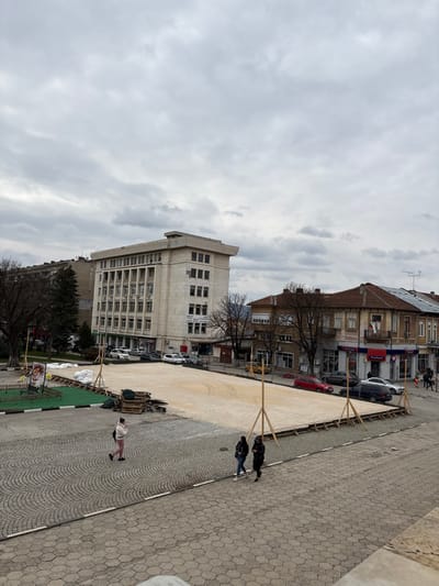 An elevated view captures a public square in Cherven Bryag, Bulgaria, on an overcast day. A large, rectangular, ground-level structure with a light-colored, possibly gravel or sand, surface occupies the midground. This structure is bordered by wooden framing and supported by several wooden tripod braces, indicating ongoing construction or temporary installation. To its immediate left, a smaller area features green turf, stacked wooden pallets, white material bags, and a display sign.

The foreground consists of a paved surface with hexagonal tiles and cobblestones. Three pedestrians are visible: one individual walks left-to-right across the cobblestones wearing light-colored pants and a light jacket, while two others walk right-to-left closer to the temporary structure, dressed in dark clothing.

In the background, a multi-story, light-colored building, identified by a banner as "ОБЩИНА ЧЕРВЕН БРЯГ" (Municipality of Cherven Bryag), stands on the left, with several cars parked in front. To the right, a row of older buildings with tiled roofs lines a street, where additional cars are parked. One building clearly displays "Пощенска Банка" (Postbank) signage and a small Bulgarian flag. Bare deciduous trees are scattered throughout the scene.