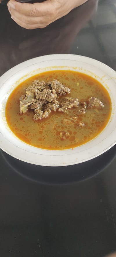 A close-up, overhead shot shows a bowl of Indonesian beef soup on a dark, reflective surface. The soup, a rich golden-brown color, contains pieces of shredded beef and appears to have a layer of oil droplets on the surface. The white bowl has a subtle grey rim. In the upper portion of the image, a person's hands are visible, one holding a small, dark object. The scene is set in Buengbakjok, Indonesia.
