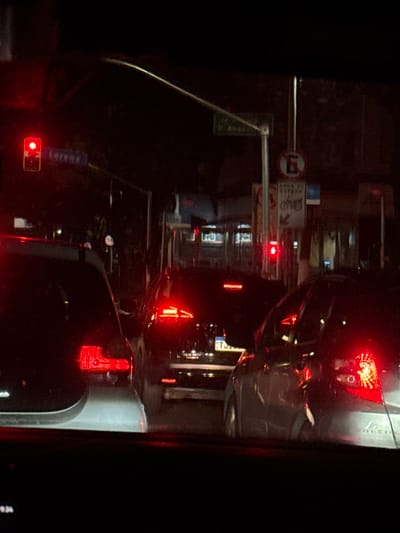 The image presents a forensic, matter-of-fact view of an urban street scene at night, observed from inside a stationary vehicle in São Paulo, Brazil. Traffic is halted, indicated by multiple illuminated red taillights from at least three vehicles ahead and two prominent red traffic lights – one on the far left and a smaller one in the center-right.

The location is identified by street signs. A blue sign on the left displays "Lorena," referring to Rua Lorena. A partially visible green sign near the center appears to indicate "P. Augusto." Another white rectangular sign clearly reads "AREA DE ESPERA" (Waiting Area) with bicycle symbols and an arrow, a common feature at intersections for two-wheeled vehicles. Above this, a red circular sign with a diagonal line through an 'E' denotes "No Parking." A small circular speed limit sign indicating "50" km/h is visible in the mid-distance.

The foreground is dominated by the dark silhouettes of vehicle bodies and their bright red brake lights. The car directly ahead on the left features bright, solid red rectangular taillights and a silver rear bumper. The central car exhibits distinct horizontal LED taillights, with a Brazilian license plate partially visible, starting with "TJ." The car on the right has intricate LED taillight patterns. The background reveals dimly lit urban buildings, suggesting commercial or residential structures. The sky is dark, with an overhead street light arc faintly visible on the upper right. No people are discernible in the scene.