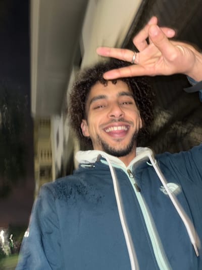 A man with curly hair smiles, making a peace sign. He wears a blue jacket, possibly wet. The background is blurred, showing what appears to be an exterior at night. Buildings with windows and dark surroundings are visible. The picture is taken at an unusual angle, seemingly rotated counter-clockwise.