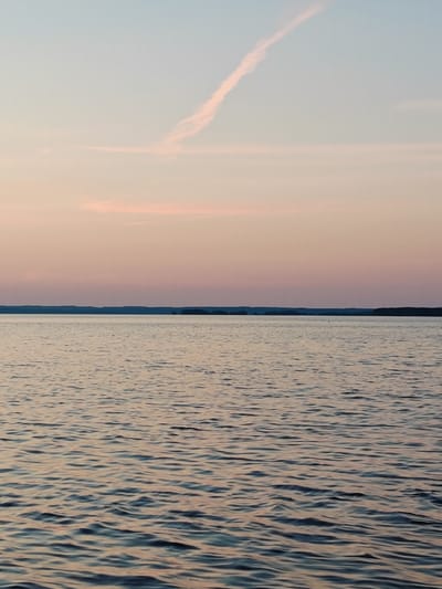 This is a photograph of a body of water, likely a reservoir or a large river, at dusk. The sky is a gradient of pale blue and soft pink, with a thin, wispy contrail visible in the upper right quadrant. A darker, tree-lined horizon separates the sky from the water. The water's surface is rippled, reflecting the muted colors of the sky and creating patterns of light and shadow. The scene evokes a sense of calm and tranquility.
The specific location in the city of Chaykovsky, Russia, is not discernible from the image itself. However, Chaykovsky is situated on the Kama River, which has been dammed to create the Votkinsk Reservoir. This reservoir is a prominent feature of the area and is consistent with the visual elements present in the photograph. Therefore, it is highly probable that this image depicts a portion of the Votkinsk Reservoir near Chaykovsky.
There are no people, animals, or man-made structures immediately visible in the foreground or mid-ground of the image. The focus is entirely on the natural landscape of the sky and water. The overall impression is one of serene natural beauty during a transitional time of day.