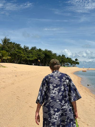 In Baan Bo Phut, Thailand, a person walks along a sandy beach. The person is viewed from behind, wearing a dark blue shirt with a white dragon and bamboo pattern, and a green baseball cap. The beach is wide and sandy, bordered by a line of lush green palm trees and other tropical vegetation. The ocean is calm and blue, with a small yellow and red boat in the distance. The sky is a clear blue with scattered white clouds. The overall scene depicts a tranquil tropical environment.