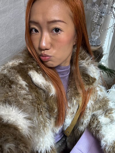 An Asian woman with reddish-brown hair lies on a white, textured surface. She wears a lilac-colored sweater and a brown and white furry coat. Her skin is dewy with blushed cheeks and glossy lips. A chandelier is visible in the upper left corner.