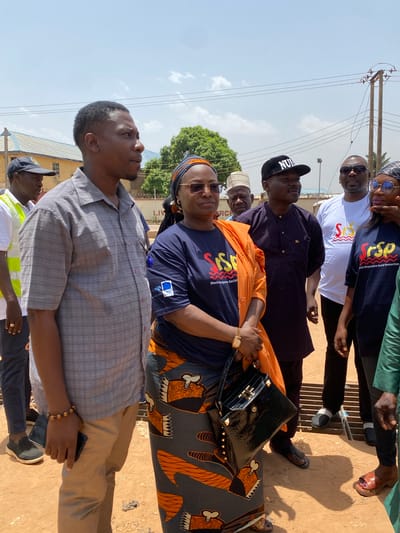 A group of people stand in Sabon Tasha, Nigeria, evident from the presence of power poles and overhead wires. The people, men and women, stand on bare ground. A woman in the center wears a navy blue top with “Shock-Responsive Social Protection” printed on it and carries a black, textured handbag. She also wears a patterned skirt. One man is wearing a hat with the word “NUIS” on it.
