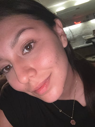 A close-up, tilted selfie of a woman with light skin, brown hair, and brown eyes. She is wearing a gold necklace with a hexagonal pendant. The background appears to be indoors, with blurred lighting and objects that are not identifiable. The location in San Andrés Cholula, Mexico, cannot be determined.