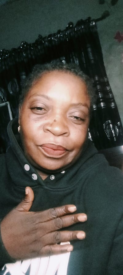 A woman with dark skin and short hair wearing a black hooded sweatshirt is the primary subject. Her hand rests on her chest. The sweatshirt has white buttons, and she wears small gold hoop earrings. Dark, shiny decorations hang behind her. The lighting is dim, contributing to a somewhat grainy quality in the image. Her lips are a muted red or brown color.