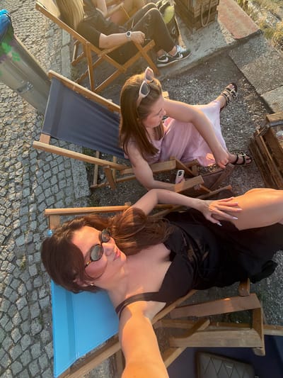 Two women are relaxing on deck chairs on a cobblestone ground, possibly in Belgrade, Serbia, given the architectural style of the surrounding elements. The woman in the foreground, wearing a black dress and sunglasses, is lying down on a blue deck chair, with her hand resting on her leg. The woman in the background, wearing a pink dress, is seated in a wooden deck chair, with her legs crossed and sandals on her feet. Both women appear to be enjoying a sunny day. The setting includes a large wooden crate and a grey bin. The lighting suggests it is late afternoon or early evening.