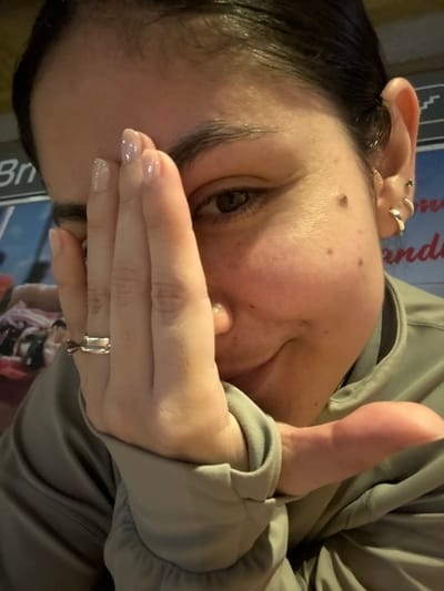 A woman with dark hair and a fair complexion is partially visible, with one eye open and looking directly at the viewer. Her hand, adorned with a silver ring, covers part of her face. She's wearing a light green hooded jacket. Her ear is visible, featuring multiple earrings. The background includes a sign with lettering. The image is rotated, presenting a tilted perspective.