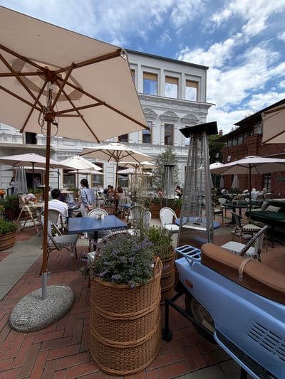 The image depicts an outdoor seating area of a cafe or restaurant, likely in Ufa, Russia. The scene is filled with tables and chairs set up on a brick-paved walkway. Several large, light-colored umbrellas provide shade over the seating. In the background, a white building with multiple windows and ornate architectural details is visible. A light blue scooter is parked on the right side of the image, with a brown leather seat. Large wicker baskets filled with plants and flowers are also placed around the seating area.
Several people are seated at the tables, some appearing to be patrons and others possibly staff. One person, possibly a waiter, is interacting with a table. Another person is pushing a stroller. The atmosphere seems relaxed and leisurely, suggesting it is daytime, likely late morning or afternoon, given the bright lighting and clear sky with some clouds. There are no overt signs of a specific event, but rather a typical day at an outdoor dining establishment.
One of the umbrellas shows a small black tag attached to its mechanism. The scooter has a red reflector and a visible license plate that is unreadable due to the resolution. A tall outdoor heater is positioned near the tables. The general ambiance is one of a pleasant, possibly warm, day, inviting people to dine outdoors.