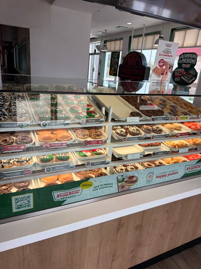 An eye-level, tilted shot captures a vibrant Krispy Kreme donut display case in San Bernardino Tlaxcalancingo, Mexico. The foreground features rows of various donuts, neatly arranged on shelves within the refrigerated display. Observable donut types include "COOKIES & KREME," "DONA GOL," "BROWNIE," "FRAMBUESA" (raspberry), "CHISPAS" (sprinkles), "DOBLE CHOCOLATE" (double chocolate), "CHOCOBROWNIE," "DONA MEX," and "minis," as indicated by individual tags. Many donuts are decorated with colorful sprinkles, chocolate drizzles, and glazes, while some appear plain or fruit-filled.

To the right of the display, a green and white "Krispy Kreme REWARDS" sign is visible, encouraging customers to "COMPRA Y ACUMULA happy points" (Buy and accumulate happy points) and displaying a QR code. Reflections on the glass of the display case show parts of the store's interior, including ceiling lights and promotional signage. One reflected sign advertises "¡ACOMPAÑA TU PASION!" (Accompany your passion!) with a football theme and mentions "TU CAJA FUTBOLITO" (your football box). Another reflection shows a sign for "A COMPARTIR una Docena La Caminera" (To share a dozen La Caminera) for $20 (likely pesos), featuring a hand holding a donut. A red "HOT ORIGINAL GLAZED" sign is also reflected. The background reveals a light-colored floor, possibly wood laminate. No people are directly visible in the frame.
