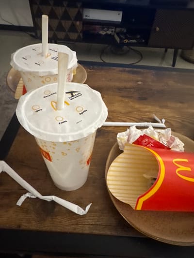 The image shows McDonald's trash on a wooden table. Two disposable cups with straws are present, one with "Sprite", "Fanta", and "Coca-Cola" printed on the lid. A McDonald's french fry container is also visible. The environment suggests an indoor setting, possibly a home or restaurant table. No people are visible. The location in the city of Cholula, Mexico, cannot be verified.