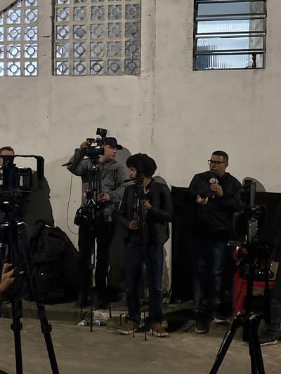 A group of people, primarily cameramen, are gathered outside a building with a window and a wall made of decorative blocks. The cameramen are operating video cameras mounted on tripods. One man in glasses appears to be giving an interview or statement while holding a microphone. This scene takes place in an urban area. It is not possible to definitively confirm the city of São Paulo, Brazil without more contextual information.