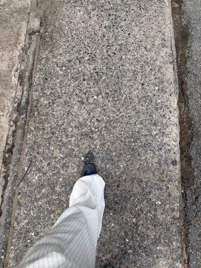 The image shows a close-up, overhead view of a person's lower leg and foot walking on a concrete sidewalk. The sidewalk is textured with small grey and black pebbles and has a raised edge. To the right of the sidewalk is a strip of darker, rougher asphalt. The person is wearing white pants with thin blue pinstripes and dark blue shoes. The scene appears to be outdoors, possibly in an urban environment, given the presence of a sidewalk and asphalt. The lighting suggests it is daytime. The location in Bangkok, Thailand, is not explicitly identifiable from the visual cues in the image.