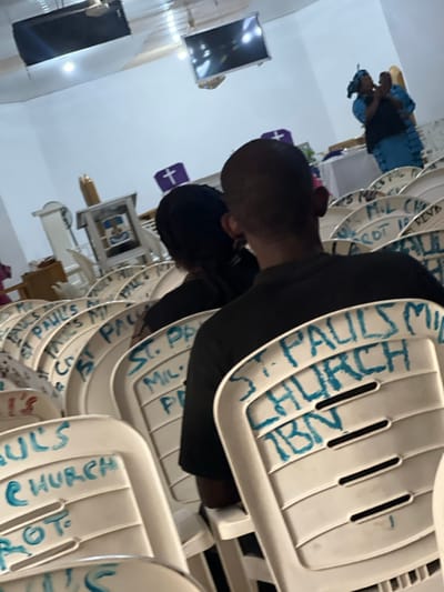 The image shows the interior of St. Paul's church. Several rows of white plastic chairs are visible, each labeled in blue marker with "St. Paul's Church." A person wearing a black shirt is seated. A woman in a blue dress is also visible in the background. The room is bright, with two TVs hanging from the ceiling and a purple cross displayed on a wall.