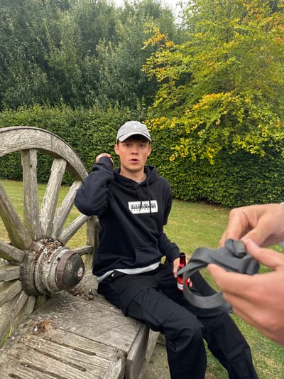 A young male is seated on a weathered wooden bench that incorporates a large wagon wheel, located within a garden environment in Bishop Wilton, United Kingdom. He wears a grey baseball cap, a black hooded sweatshirt with a white Napapijri logo, and black cargo pants. His mouth is slightly open, and he is looking directly towards the viewer; his right hand is positioned near his head. A red-labeled bottle rests on the bench between his legs. In the lower right foreground, two hands are visible, manipulating a black, flexible object with a looped section. The background consists of dense green hedges and trees, some displaying yellowish-green autumnal foliage, above a manicured green lawn.