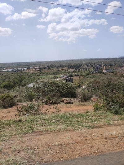 The image captures a wide, sunlit landscape view of what appears to be a rural or semi-rural area, consistent with Ewuaso Oonkidong'i ward, Kenya. The scene unfolds under a bright blue sky dotted with scattered white cumulus clouds.
In the foreground, a reddish-brown dirt path or ground stretches across the bottom, featuring sparse patches of green and dry grass and low-lying bushes. A dark, possibly asphalt, surface or car window ledge is visible at the very bottom edge of the frame.
The midground and background are dominated by an expansive, undulating terrain covered in dense natural vegetation, comprising a mix of green and dry bushes and trees, characteristic of a bushland or savannah environment. Scattered within this natural expanse are various buildings and small settlements. Some structures appear to be complete houses, while others, particularly in the midground, show exposed brickwork or concrete, suggesting ongoing construction or unfinished development. These buildings are spread out, indicating a low-density population.
Overhead, power lines are visible, stretching horizontally across the sky from left to right. In the far distance on the right, several tall electricity pylons or transmission towers stand against the horizon, indicating established infrastructure.
The time of day is clearly daylight, likely late morning or early afternoon, given the bright illumination and visible cloud patterns. No human activity is discernible, and no text is visible within the image. The overall impression is a vast, natural landscape with evidence of gradual human settlement and development.