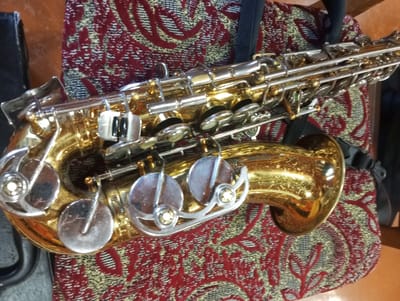 This is a medium-angle, close-up shot of a saxophone resting on a patterned fabric. The saxophone is the primary subject, gleaming with a golden finish and accented with silver keys and mechanisms. The fabric it rests on has a rich, intricate design in shades of red, gold, and black, suggesting a decorative cushion or upholstery.
The saxophone appears to be a tenor or alto saxophone, judging by its size and the curved bell. Key details visible include the intricate keywork, the tone holes, and the metal pads. The instrument's polished surface reflects the surrounding light, highlighting its craftsmanship.
The scene is set indoors, indicated by the lighting and the material of the surface the saxophone is placed upon. There are no people visible in the image. The overall mood is still and quiet, suggesting the instrument is not currently being played. The context provided places the scene in Oleksandriia, Ukraine, but there are no visual cues within the image itself to confirm this specific location. The image focuses solely on the instrument and its immediate surroundings. No text is visible in the image.