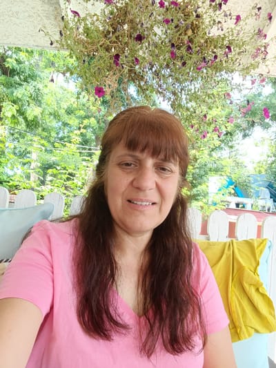 A woman with brown hair and bangs smiles at the camera, wearing a pink t-shirt. She is seated outdoors on what appears to be a porch or patio, with a white picket fence behind her. Above her head, a hanging plant with wilting purple flowers is visible, suggesting it might be late summer or early autumn. Green foliage from trees can be seen in the background, indicating a natural setting. A bright yellow garment is draped over a chair or railing to her right, and a light blue pillow is visible to her left. The lighting suggests it is daytime, possibly late morning or afternoon, with soft sunlight filtering through the leaves. The overall atmosphere is relaxed and casual. The location is identified as Harmanli, Bulgaria.