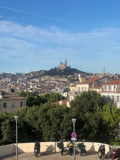 The image captures a scenic view of Marseille, France, dominated by the iconic Notre-Dame de la Garde basilica perched atop a hill. The city sprawls across the landscape, a dense collection of buildings with terracotta roofs, extending towards the horizon under a clear blue sky dotted with wispy clouds. In the foreground, several scooters are parked along a paved area bordered by a low, cream-colored wall. Green trees provide a lush backdrop, partially obscuring some of the buildings. The lighting suggests it is either early morning or late afternoon, with shadows cast by the parked scooters and streetlights.