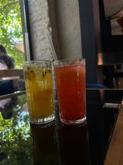 Two glasses of brightly colored drinks sit on a reflective surface, likely a table, in what appears to be a casual setting with outdoor elements visible in the reflection. The top glass contains a reddish-pink drink with some pulp or fruit pieces. A clear plastic straw extends from this glass. Below it, another glass holds a vibrant yellow drink, also with visible seeds or fruit matter, and a clear plastic straw. The drinks are tall and appear to be refreshments. The reflective surface shows a blurred background of greenery and light, suggesting it might be daytime and the location is near trees or foliage. The overall impression is one of a relaxed moment, possibly at a cafe or outdoor dining area. The background also shows some indoor elements and furniture, indicating a transition between indoor and outdoor spaces. No visible text is discernible in the image.