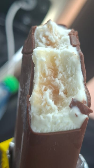 A close-up shot captures a partially eaten ice cream bar with a chocolate coating. The ice cream inside is white with a swirl of caramel or golden-colored syrup. The chocolate shell is slightly broken, revealing the creamy interior. The background is blurred and dark, with hints of light and color, possibly indicating an indoor or vehicle setting. No specific location in Лугаваслабадскі сельскі Савет, Belarus can be determined from the image.