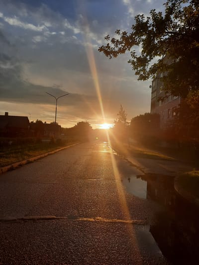 The image depicts a street scene in Paretcha, Belarus, during a sunset. The sun is low on the horizon, casting a strong, golden light and creating visible sun rays. The asphalt road is wet, with puddles reflecting the surrounding environment. On the left side of the road, there are several houses and trees. On the right side, a multi-story building is visible, partially obscured by trees. The sky is a mix of clouds and blue sky, with dramatic lighting from the setting sun. There are no people visible in the image. The overall impression is one of a quiet, residential street at the end of the day.