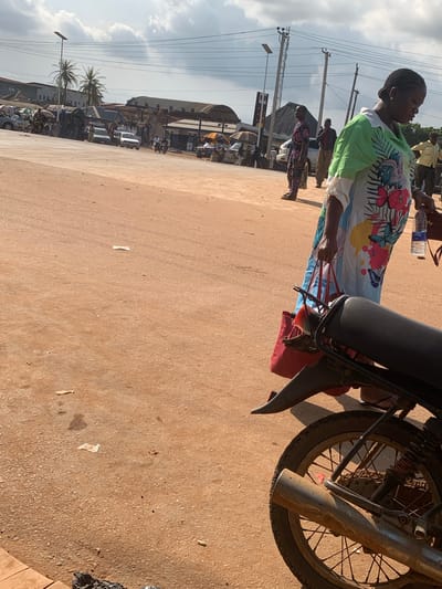 A person is standing near a motorcycle in a street scene in Shagamu, Nigeria. The person is wearing a colorful, patterned dress and has their hair in braids. They are holding onto the back of the motorcycle, which has a dark seat and a spoked wheel. In the background, there are other people, buildings, and vehicles, including cars and more motorcycles. Power lines and utility poles are visible against a cloudy sky, suggesting daytime. The ground appears to be a dusty, reddish-brown dirt or gravel road. The overall impression is of a busy street or market area.