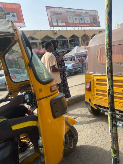 A sunny outdoor scene features yellow auto rickshaws. One is in the foreground, with a person wearing black and yellow pants appearing to work on it. Another rickshaw is visible behind, partially obscured. Two individuals stand nearby, one holding a phone. A building lines the street with advertising billboards, including one promoting solar panels, batteries, and inverters. A street market or event appears in the distance.