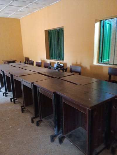 This image shows an interior view of an empty classroom in Kakuri Gwari, Nigeria. The room has an orange-yellow painted wall and a tiled ceiling. There are two windows, both with green, slatted shutters. The room is filled with rows of dark wooden desks, each with a cubbyhole underneath. The desks are arranged in long tables, suggesting they are for a lecture hall or a large classroom setting. Several wooden chairs are visible behind the desks, some pushed in and others pulled out. The lighting appears to be natural, coming from the windows, and suggests it is daytime. The floor is a concrete material. There is no text visible in the image. The scene is devoid of people, creating a quiet and potentially abandoned atmosphere.