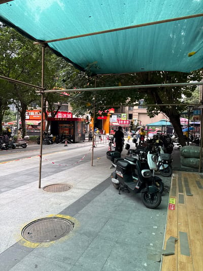 The image depicts a street scene in Haizhu District, China, likely a commercial area, under a large teal-colored tarp supported by scaffolding. In the foreground, several electric scooters are parked along a sidewalk. One scooter in the immediate foreground is black with orange accents and the number "05" visible. To its right, a white scooter is parked. Further back, more scooters are lined up. The sidewalk is paved with gray tiles, and two manhole covers are visible. One manhole cover in the foreground is surrounded by yellow spray paint. A red and white striped tape barrier is stretched across the sidewalk in the middle ground.
Across the street, there are storefronts. One red and black structure with Chinese characters that appear to be a food or drink vendor is visible. Behind it, other buildings with storefronts and awnings can be seen. People are present on the street, including a person in a yellow top and a dark-suited person walking away from the camera. Trees with green foliage are scattered throughout the scene, providing shade. The overall impression is a busy urban street with a temporary structure overhead, possibly for shade or construction.