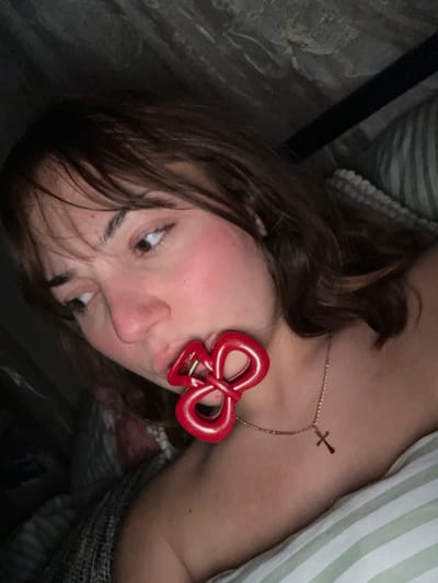 A woman lies on her side in bed, facing the camera. She has brown hair and her face is close to the lens. A red bow-shaped hair clip is attached to her eyebrow. She is also wearing a delicate gold necklace with a small cross pendant. The bedding includes a striped pillow and a floral-patterned blanket. The overall lighting is subdued, and the image is slightly rotated.