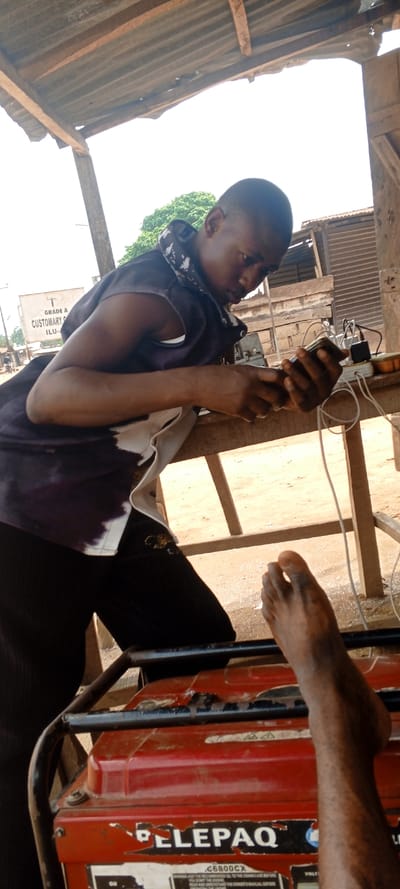 The image depicts an outdoor, partially sheltered scene in Ago-Abo, Nigeria. A young Black male with short hair, wearing a dark sleeveless top over a white and patterned shirt and dark trousers, is positioned centrally, leaning forward over a wooden table. He holds a mobile phone in both hands, which is connected by a white cable to a white multi-socket power strip on the table. Several black power adapters are plugged into the power strip. In the lower foreground, a red "ELEPAQ C6800CX" portable generator with a black metal frame is visible. A bare, dark-skinned human foot rests on the generator's frame in the lower right. The overhead structure features a corrugated metal roof supported by wooden beams. The background on the left includes a white sign with partial black text reading "GRADE A CUSTOMARY ILU-", distant green trees, and building structures. The ground throughout the visible area is dry earth.