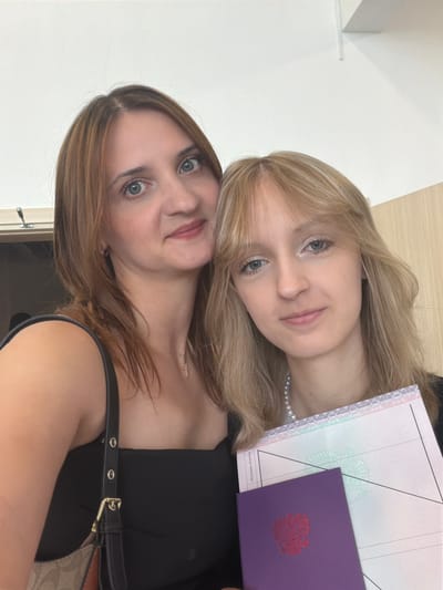 In Mytishchi, Russia, two women are captured in a selfie taken indoors. The woman on the left, with shoulder-length brown hair and blue eyes, wears a black top and a necklace. She is holding a purse with a patterned strap. The woman on the right, with blonde hair and blue eyes, is also wearing a black top and a pearl necklace. She is holding two documents: a purple one with a red emblem and a white document with intricate designs and text. The background appears to be a wall, possibly in an educational or administrative building, with a wooden texture visible on the right side.