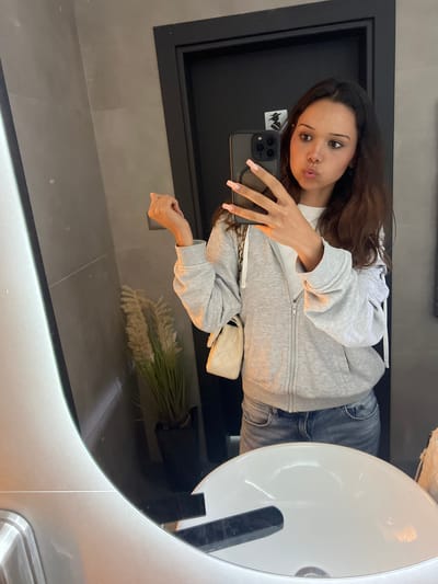 In Zlatograd, Bulgaria, a young woman with brown hair and light skin is taking a mirror selfie in what appears to be a public restroom. She is wearing a grey zip-up hoodie over a white t-shirt and blue jeans. Her nails are painted a light pink. She is holding a smartphone in her right hand, with her left hand resting on the side of the mirror. In the background, a potted plant with dried grass-like stalks is visible, along with a dark door and grey tiled walls. A white purse with a quilted texture is hanging from her left shoulder. The sink in the foreground is white and modern in design.