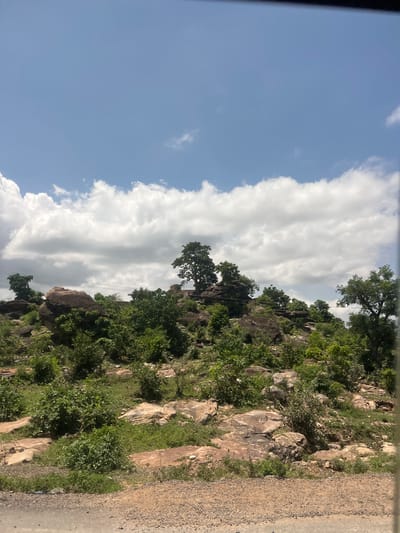 This image displays a natural, rocky hillside landscape under a partly cloudy blue sky in Lafia, Nigeria. The upper portion of the frame features a clear blue sky interspersed with large, white cumulus clouds. The terrain consists of an upward-sloping expanse of numerous dark brown and reddish-brown boulders and exposed rock formations. Patches of green shrubbery and trees are densely scattered across the rocky ground, with several larger trees prominent on the higher elevations. Multiple thin, dark power or communication lines stretch horizontally across the mid-ground, passing above the vegetation and rocks. The extreme bottom of the image reveals a narrow strip of a dirt or gravel road, suggesting a roadside viewpoint. No people or animals are visible in the scene.