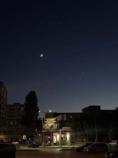 The image shows a nighttime scene in the city of Shumen, Bulgaria. In the sky, a crescent moon is visible along with several bright stars or planets. Below, the silhouette of buildings and trees is apparent. A street light illuminates a billboard advertising residential buildings and a store or food stand. Several cars are parked in the foreground and middle ground. There are no people clearly visible in the scene, but there appear to be a few figures in the distance near the store.