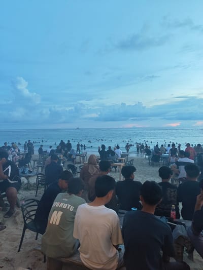 The image shows a beach scene at Monikeun, Indonesia. A large group of people are visible both in the water and on the sand. Tables and chairs are set up on the beach, and groups of people are seated, facing the ocean. Some individuals are wading or swimming in the sea. The sky is a pale blue with scattered clouds, suggesting either dawn or dusk. The overall scene suggests a casual social gathering at the beach.