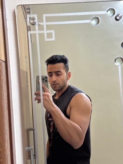 A dark-haired man in a sleeveless black shirt is taking a mirror selfie. His muscular arm is visible, holding a silver smartphone with a triple-lens camera. The mirror has a decorative pattern with silver knobs and linear etchings. The background is a light cream color. A wooden surface is visible below the mirror's edge.