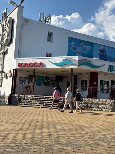 The image shows the exterior of a building with a sign that reads "KACCA", which is Russian for "ticket office". There are several people walking on a paved walkway in front of the building. The building appears to be a tourist attraction, possibly a dolphinarium, as suggested by the large mural of dolphins on the facade. The weather is sunny with some clouds in the sky. The overall scene depicts a typical day at a public venue in Gelendzhik, Russia.