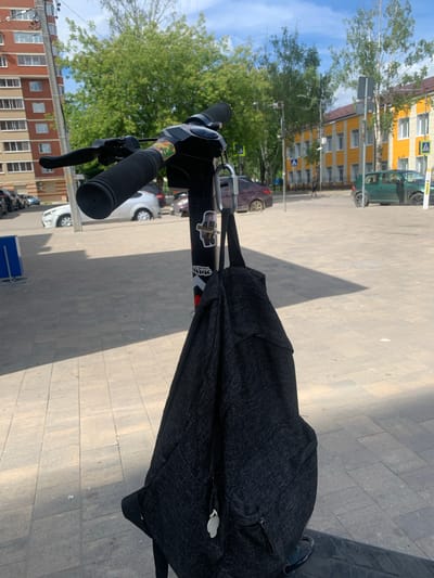 An electric scooter is parked outdoors on a paved area. The scooter's handlebars are visible in the foreground, with a black backpack hanging from the handlebars by a carabiner. The backpack is dark gray and appears to be made of a textured fabric. The scooter has a black stem, and there are two stickers on it. One sticker features a cartoon character, and another has a geometric design. In the background, there are several buildings, including a multi-story brick apartment building and a yellow building with a red roof. Trees line the street, and several cars are parked in the area. The sky is blue with some scattered clouds. The image appears to have been taken in the city of Klin, Russia.