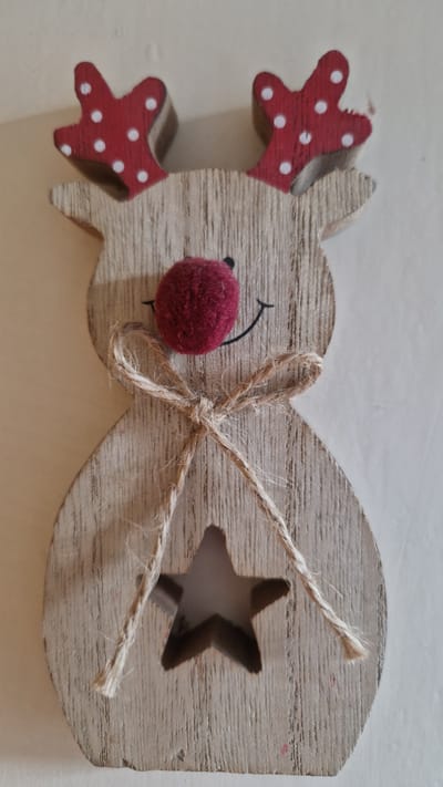 A wooden reindeer decoration is pictured against a plain background. The reindeer is made of light-colored wood with a visible grain. It has red antlers with white polka dots, a dark red pom-pom nose, and a simple black smile. A twine bow is tied around its neck. A star-shaped cutout is present in the lower portion of the reindeer's body. The image is a close-up, top-down shot of the object. There is no indication of its location in Riga, Latvia.