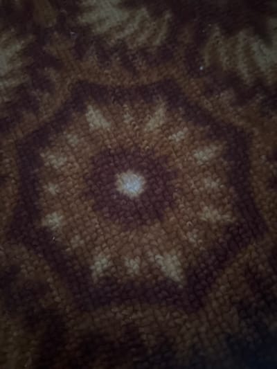 The image displays a close-up, blurry view of a textured surface, likely fabric, with a patterned design. The pattern appears to be a repeating geometric motif, possibly a star or floral shape, rendered in shades of brown and tan. The texture of the material suggests it could be a blanket, rug, or upholstery. The lighting is dim, creating deep shadows and highlighting the textured surface. There are no discernible people, objects, or specific setting details visible. No activity or event is taking place, and there is no visible text. The overall impression is abstract and tactile, focusing on the texture and pattern of the material. The context of Kirov, Russia, does not provide any specific visual cues within the image itself.