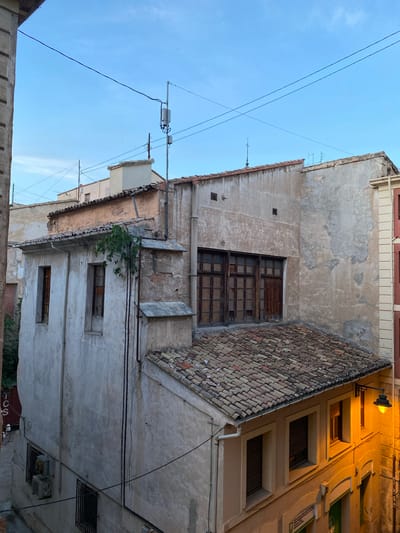 This is a vertical, low-angle shot of the exteriors of several old, weathered buildings in a narrow street in Alcoy, Spain. The sky is a clear, pale blue, indicating daytime. The buildings have stuccoed walls, some showing signs of peeling paint and age, with red-tiled roofs.
One building in the foreground has multiple windows, some with wooden frames and glass panes, and one larger window that appears to be boarded up or heavily damaged. A small tree or vine grows out of a window ledge. A tiled roof juts out at an angle, casting a shadow. Electrical wires crisscross the sky, and a street lamp with an orange glow is visible on the side of a building to the right, suggesting it might be dusk or dawn, or that the street is dimly lit.
Another building to the right features several rectangular windows set into recessed frames, illuminated by the warm glow of the street lamp. The overall impression is one of urban decay and older architecture. There is no visible text in the image.