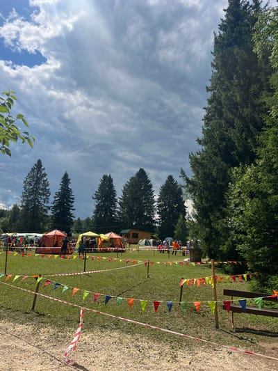 The image depicts an outdoor event, possibly a festival or gathering, set in a grassy field with a backdrop of tall evergreen trees. The sky is partly cloudy, suggesting it is daytime. Several tents of various colors, including orange, yellow, and green, are pitched on the field, indicating temporary accommodation or vendor stalls. A row of small, colorful flags—orange, yellow, green, and blue—are strung along poles, marking a boundary or path.
In the foreground, a gravel path runs alongside the field, separated by a red and white striped tape. People are visible in and around the tents, suggesting active participation in the event. The overall scene conveys a festive and lively atmosphere. The location is identified as Sergino, Russia, and the time of day appears to be sunny. There is no visible text in the image.